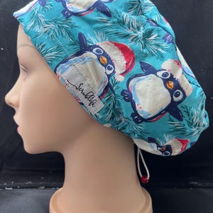 May include: A teal scrub cap with a pattern of penguins in Santa hats and pine sprigs. The cap has a white tag with the text "Scrublife". The cap is displayed on a mannequin head.
