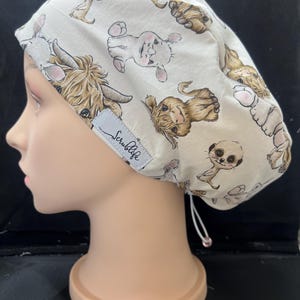 May include: A cream-coloured scrub cap with a repeating pattern of cartoon Highland cows and meerkats in shades of brown and tan. The cap has a drawstring and a small label that reads "Scrublife".