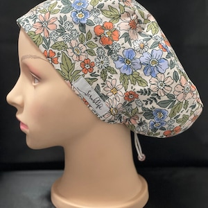 May include: A white scrub cap with a floral print featuring pink, blue, and orange flowers. The cap has a tie at the back and the word "Scrublife" is printed on the side.