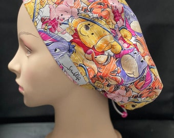 Könnte beinhalten: Eine bunte OP-Mütze mit Winnie Puuh und Tigger-Motiv. Die Mütze hat einen Bindeverschluss an der Rückseite und der Schriftzug "ScrubLife" ist seitlich aufgedruckt.
