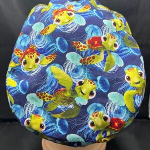 May include: A navy blue surgical scrub cap featuring a repeating pattern of cartoon sea turtles and jellyfish. The turtles are yellow and green with red accents, and the jellyfish are blue and white. The cap is designed to cover the head.