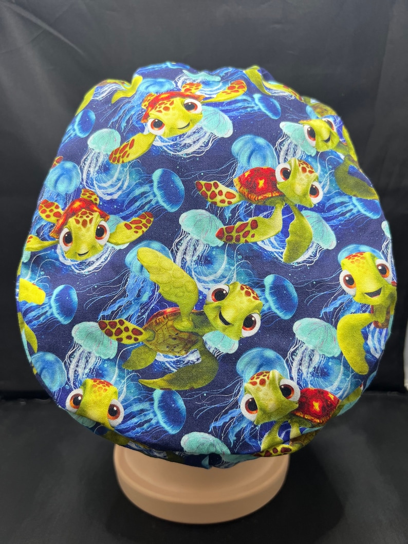 May include: A navy blue surgical scrub cap featuring a repeating pattern of cartoon sea turtles and jellyfish. The turtles are yellow and green with red accents, and the jellyfish are blue and white. The cap is designed to cover the head.