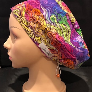 May include: A colourful surgical scrub hat with a rainbow swirl pattern. The hat has a white label with the text "Scrublife" on it. The hat is tied with a white drawstring and a small orange bead.