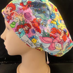 May include: A brightly patterned scrub cap with illustrations of a mermaid, a red crab, and other marine life. The cap is made of a soft fabric and has a drawstring for an adjustable fit. The text "Scrublife" is visible on the side.