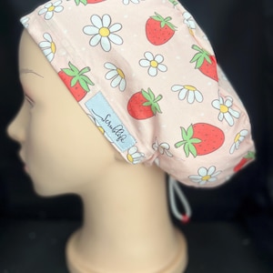 May include: A light pink scrub cap featuring a repeating pattern of red strawberries and white daisies with yellow centers. The cap includes a light blue rectangular fabric tag with the text Scrublife in black script and a drawstring closure.