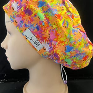 May include: A brightly coloured scrub cap with a pattern of cartoon trolls in orange, yellow, pink, and blue. The cap has a white label that reads "Scrublife". The cap is on a mannequin head.