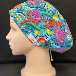 May include: A patterned scrub cap in teal, pink, orange, and yellow floral design. The cap has a white tag with the text "Scrublife". The cap is designed for medical professionals. The cap is designed to be worn in a medical setting.