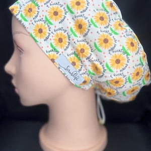 May include: A white scrub cap featuring a repeating pattern of yellow sunflowers with green stems and small red hearts. The fabric is printed with the repeating text "I fucking hate people" in black. A light blue tag reads "Scrublife" in script.