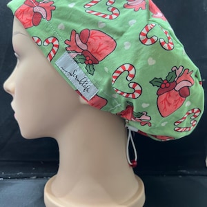 May include: A green scrub cap featuring a pattern of red anatomical hearts, candy canes, and white hearts. The cap has a white tag with the text "Scrublife". The cap is on a mannequin head.