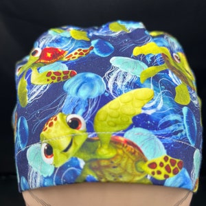 May include: A navy blue scrub cap featuring a colorful underwater scene. The cap is patterned with cartoon sea turtles, jellyfish, and other marine life in shades of green, yellow, blue, and red. The cap is designed for medical professionals.