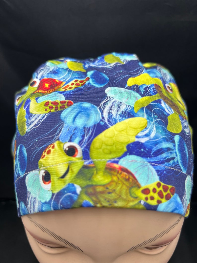 May include: A navy blue scrub cap featuring a colorful underwater scene. The cap is patterned with cartoon sea turtles, jellyfish, and other marine life in shades of green, yellow, blue, and red. The cap is designed for medical professionals.