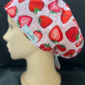 May include: A light pink scrub cap with a repeating pattern of red strawberries and green leaves. The cap has a light blue tag with the text "Scrublife". The cap is displayed on a mannequin head.