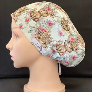 Pode incluir: Um gorro de cirurgia verde com um padrão floral com rosas cor-de-rosa e ilustrações de vacas das terras altas marrons. O gorro tem uma etiqueta branca com o texto "ScrubLife".