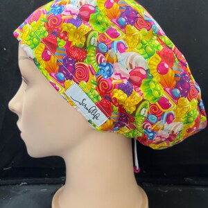 May include: A brightly patterned scrub cap with a design of sweets, lollipops, and fruit pieces in red, yellow, green, and purple. The cap has a white tag with the text "Scrublife".
