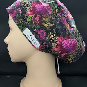Peut inclure: Un bonnet de chirurgien à motifs floraux avec un fond noir, un motif en nid d'abeille doré et des fleurs roses et violettes. Le bonnet porte une étiquette blanche avec le texte "Scrublife". Un cordon de serrage blanc est visible.