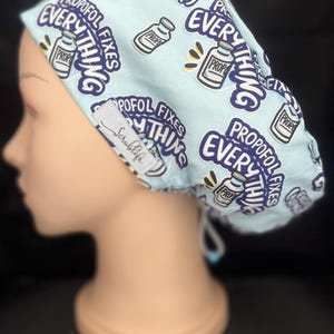May include: A light blue scrub cap featuring a repeating pattern of medical vials and the text "Propofol fixes everything" in dark purple lettering. A small white fabric tag with the brand name "Scrublife" is stitched onto the side of the cap.