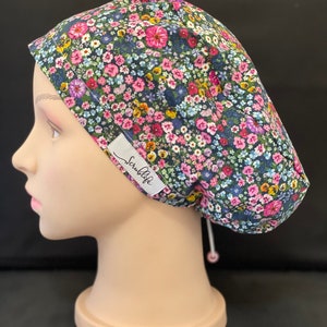 May include: A dark blue scrub cap with a floral print featuring pink, yellow, and white flowers. The cap has a drawstring with a small bead at the end. The brand name "Scruffy" is printed on the side of the cap.