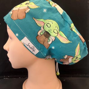 Puede incluir: Gorro quirúrgico de color turquesa con un patrón repetido de un personaje alienígena verde con orejas grandes y túnicas marrones. La gorra tiene una etiqueta blanca que dice "Scrublife". Se ve un cordón blanco.