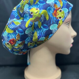 May include: A blue scrub cap featuring a pattern of cartoon sea turtles and jellyfish. The turtles are green, brown, and yellow, while the jellyfish are blue and white. The cap is tied at the back with a white string.