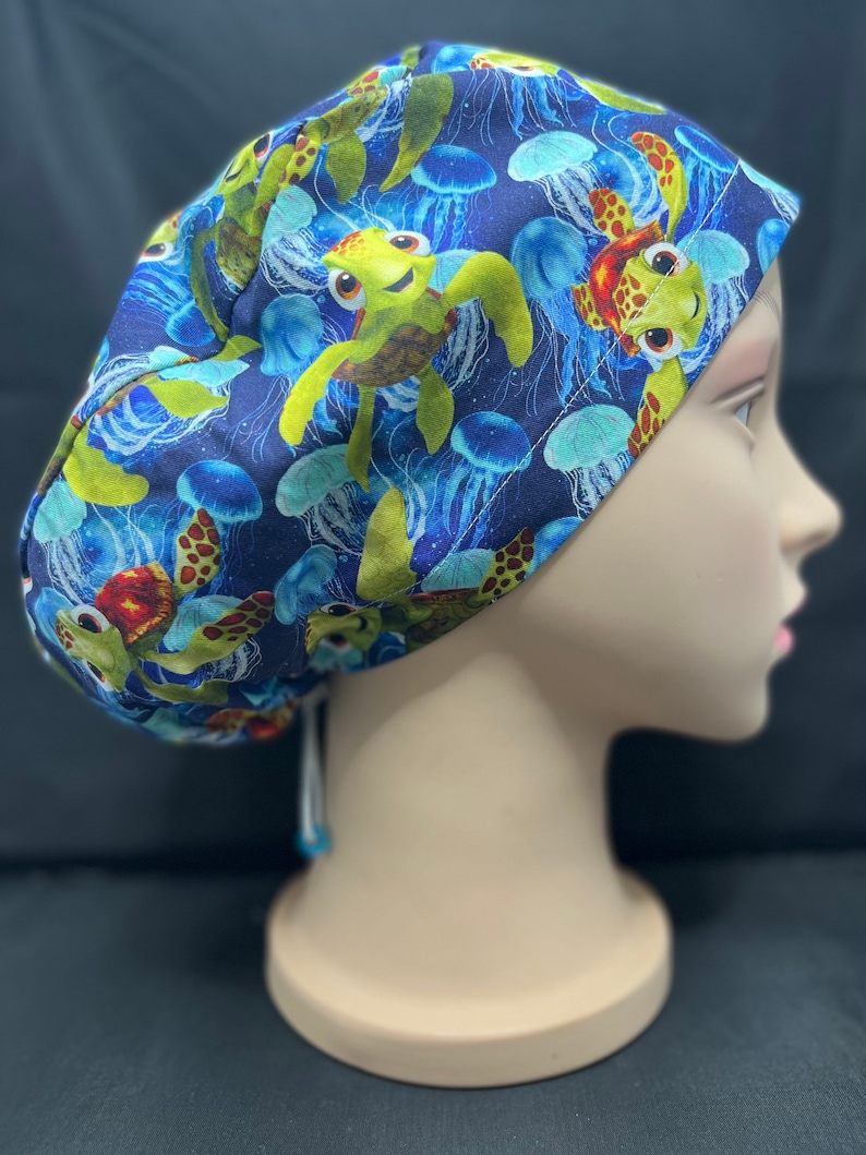 May include: A blue scrub cap featuring a pattern of cartoon sea turtles and jellyfish. The turtles are green, brown, and yellow, while the jellyfish are blue and white. The cap is tied at the back with a white string.