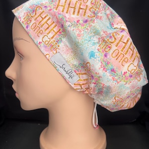 May include: A scrub cap featuring a light blue and pink watercolour background with floral accents and gold glitter text that reads SHHH no one cares. The cap includes a white adjustable drawstring at the back and a grey label with the text Scrublife.