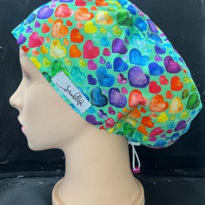 May include: A scrub cap with a vibrant heart pattern in shades of red, orange, yellow, green, blue, and purple, set against a light green backdrop. The cap includes a white label with the text "Scrublife".