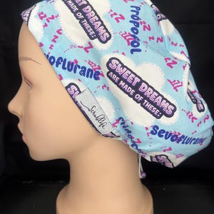 May include: A light blue scrub cap featuring a repeating pattern of white clouds, pink Zzz symbols, and the text Sweet Dreams Are Made Of These, Propofol, Sevoflurane, and Isoflurane. A small grey tag with the text Scrublife is attached to the side.