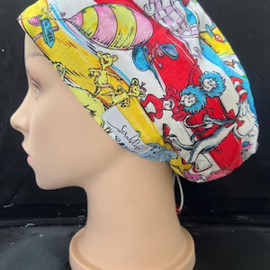 May include: A colorful surgical scrub cap featuring a whimsical design with characters and imagery from a children's book. The cap is predominantly white, red, yellow, and blue, with the text "To There" visible. The cap has a tag that says "Scrubble".