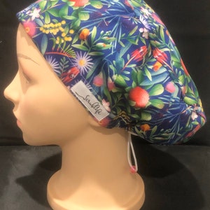 Può includere: Un berretto chirurgico blu con una stampa floreale con fiori rosa, rossi e gialli. Il berretto ha un cordoncino bianco e un'etichetta bianca con la scritta "ScrubLife".