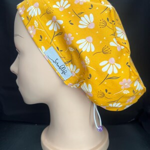 May include: A mustard yellow scrub cap featuring a repeating pattern of white and light pink daisies with small black stems and star accents. The cap includes a light blue rectangular tag with the text Scrublife and a white elastic cord with a purple bead.