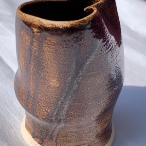 Puede incluir: Una pequeña jarra de cerámica marrón con un borde blanco y una forma única y asimétrica. La jarra tiene un acabado brillante y un patrón moteado sutil.