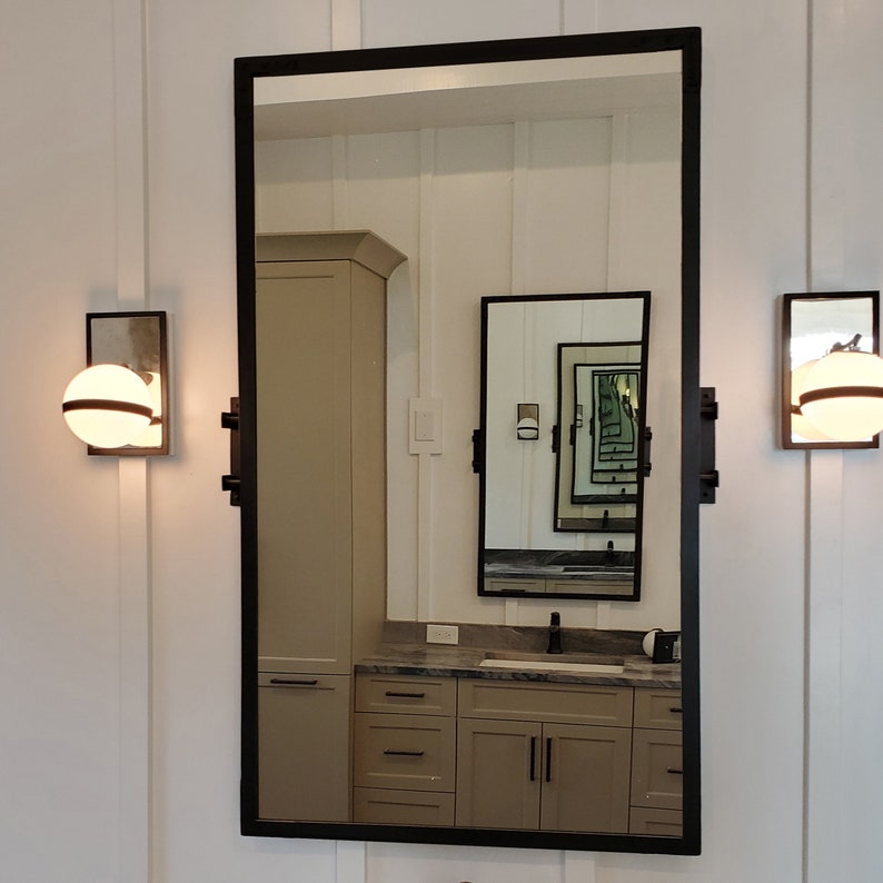 May include: A black framed rectangular mirror with a white wall and a vanity with a sink and cabinets reflected in the mirror.
