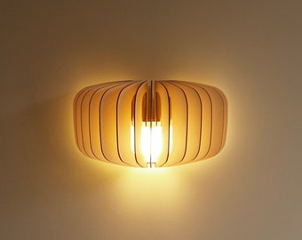 Puede incluir: Un aplique de pared construido con listones de madera de color claro dispuestos en una forma ovalada horizontal y redondeada. El diseño presenta paneles de madera verticales que permiten que una luz cálida y brillante emane del centro.