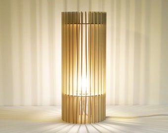 Può includere: Una lampada da tavolo cilindrica costruita con listelli di legno chiaro verticali disposti in un motivo circolare. La lampada presenta una fonte di luce interna che illumina i listelli, proiettando lunghe ombre lineari verso l'esterno.