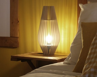 Può includere: Una lampada da tavolo con una struttura in legno chiaro composta da listelli verticali disposti in una forma ovale allungata e affusolata. La lampada emette una luce calda da una lampadina centrale, proiettando ombre soffuse attraverso i listelli.