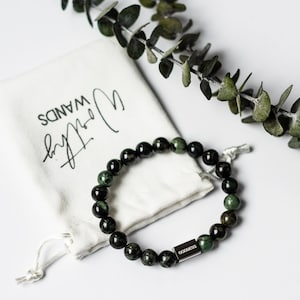 Op de afbeelding: Een groene en zwarte kralen armband met een zilveren bedel met de tekst "Godin". De armband ligt op een witte stoffen tas met de tekst "Wands by Noelle" erop.