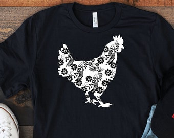 Pode incluir: Camiseta preta com um desenho floral branco de galinha. A imagem também mostra jeans azul claro, tênis brancos e um boné preto com um quadrado vermelho. Os itens estão dispostos em uma superfície de madeira.