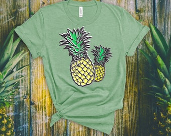 Pode incluir: Uma camiseta verde com um gráfico de dois abacaxis amarelos com topos verdes. Os abacaxis são estilizados e têm contornos pretos.