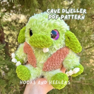 May include: A green crocheted cave dweller plush toy with a pink belly and white teeth. The toy has a single purple eye with a black pupil. The text "Cave Dweller PDF Pattern" is above the toy and "Hooks and Heelers" is below the toy.