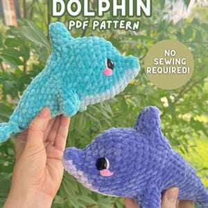 Op de afbeelding: Twee pluche dolfijnspeeltjes gemaakt van zacht, getextureerd garen. De ene is lichtblauw en de andere is paars. Beide hebben zwarte plastic ogen en roze geborduurde wangen. De tekst luidt: DOLPHIN PDF PATTERN, NO SEWING REQUIRED!, HOOKS AND HEELERS.