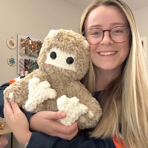 May include: A woman is holding a brown and white plush toy with black eyes. The toy has a fluffy texture and white accents on its arms and legs.