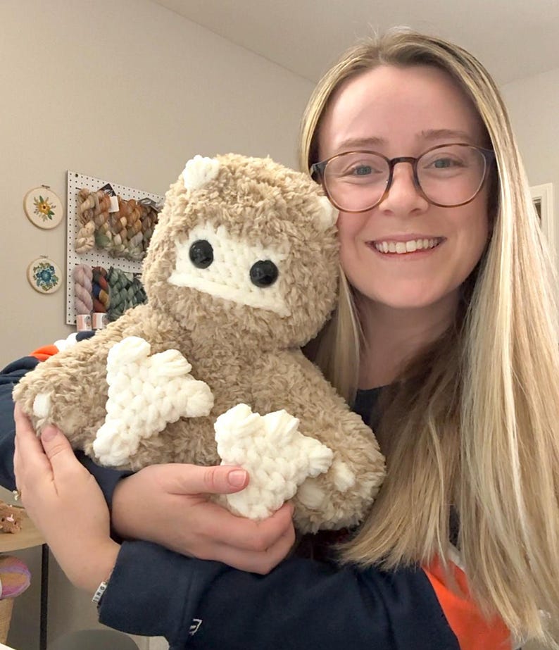 May include: A woman is holding a brown and white plush toy with black eyes. The toy has a fluffy texture and white accents on its arms and legs.