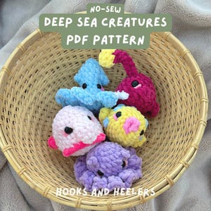 May include: A basket filled with crocheted deep sea creatures. The creatures are a pink blobfish, a yellow anglerfish, a purple octopus, a blue anglerfish, and a red anglerfish. The basket is made of woven bamboo. The text "NO-SEW DEEP SEA CREATURES PDF PATTERN" is written in green on a white background. The text "HOOKS AND HEELERS" is written in white on a black background.