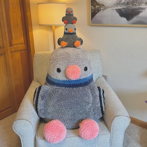 Peut inclure: Un grand coussin en forme d'oiseau en peluche grise avec un bec et des pieds roses, et un col rayé bleu et noir. Deux petits oiseaux en peluche sont posés dessus. Les coussins sont sur un fauteuil gris.