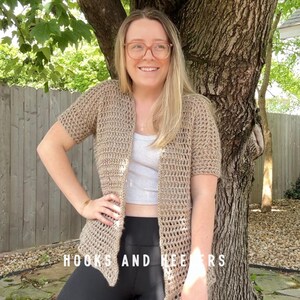 May include: A light brown, short-sleeved crochet cardigan. The open-front design features a textured stitch pattern. The text "HOOKS AND HEERS" is visible at the bottom. The cardigan is worn over a light-colored top and black leggings.