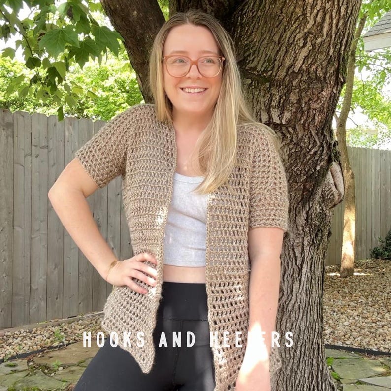 May include: A light brown, short-sleeved crochet cardigan. The open-front design features a textured stitch pattern. The text "HOOKS AND HEERS" is visible at the bottom. The cardigan is worn over a light-colored top and black leggings.
