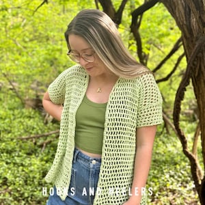 May include: A light green, open-weave crochet cardigan with short sleeves. The cardigan is worn over a matching green crop top and blue jeans. The text "HOOKS AND NEEDLERS" is visible at the bottom of the image. The background is a blurred green foliage.