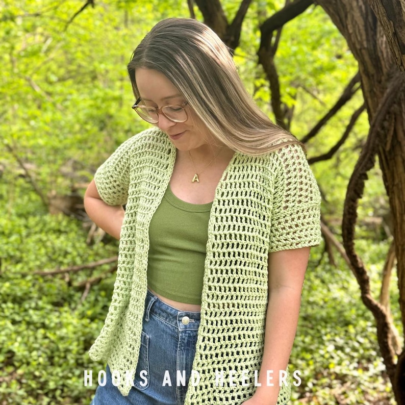 May include: A light green, open-weave crochet cardigan with short sleeves. The cardigan is worn over a matching green crop top and blue jeans. The text "HOOKS AND NEEDLERS" is visible at the bottom of the image. The background is a blurred green foliage.