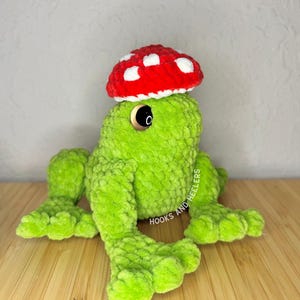 May include: A handmade green crochet frog wearing a red mushroom hat with white spots. The frog has large black eyes and is lying on a wooden surface. The text "HOOKS AND HEELERS" is visible on the side.