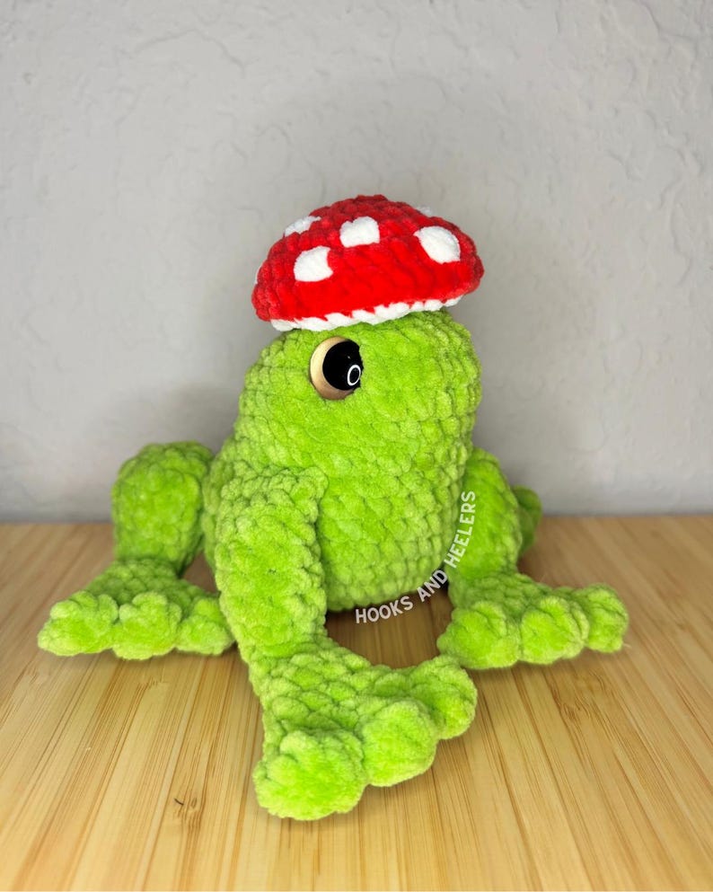May include: A handmade green crochet frog wearing a red mushroom hat with white spots. The frog has large black eyes and is lying on a wooden surface. The text "HOOKS AND HEELERS" is visible on the side.